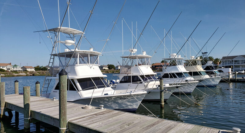 A row of sportfishing charter boats tied along a wooden pier in a small harbor, masts and outriggers against a blue sky.